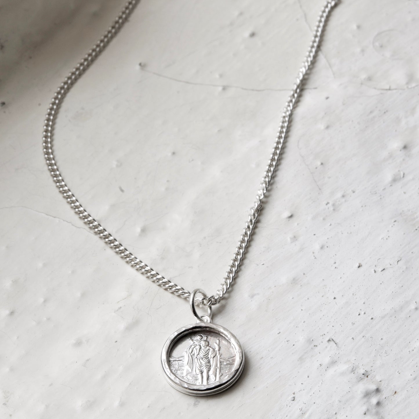 Handmade sterling silver St. Christopher with textured frame, suspended from a silver curb chain. Pictured on a rustic white back ground in the Cotswold's.